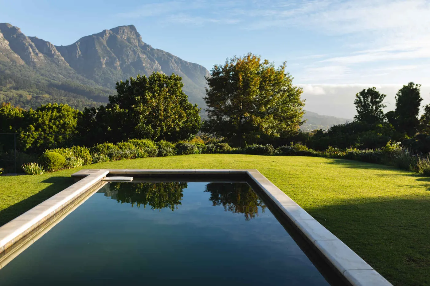 A rectangular backyard swimming pool with still, dark water, bordered by stone coping and a wide mown lawn, with trees and a mountain ridge behind it in early morning light