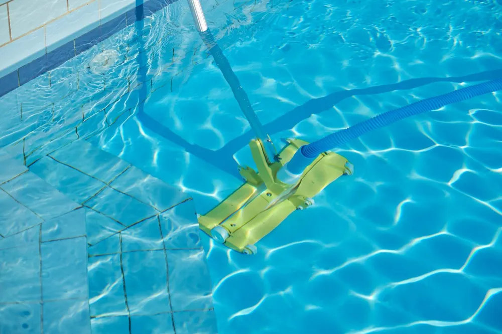 A yellow manual pool vacuum head on a telescopic pole, connected to a ribbed suction hose, resting on the tiled floor of a swimming pool