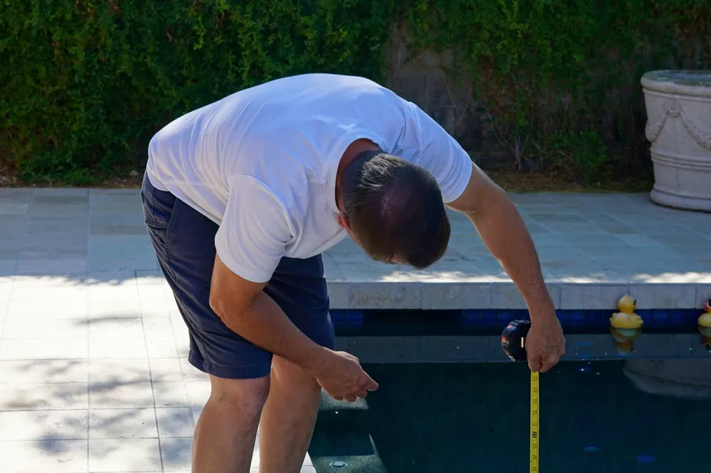 A person leaning over the edge of a swimming pool and running a tape measure down into the water to read the depth