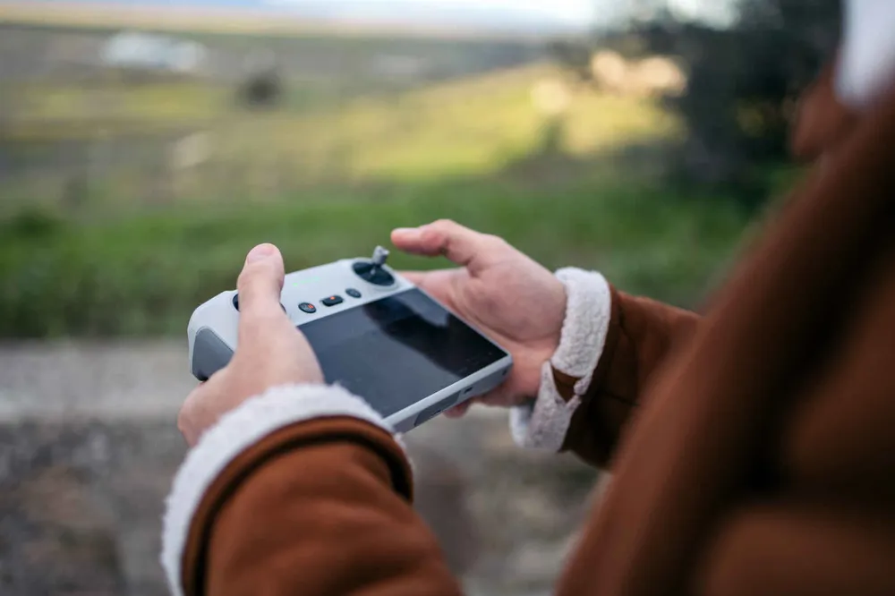 Two hands holding a handheld remote control transmitter with a screen and thumbsticks, while standing on level ground overlooking a green field