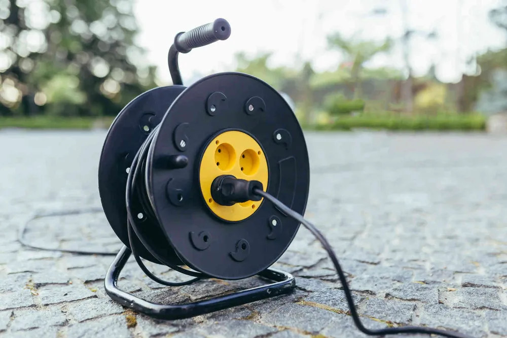 A black extension cord reel standing on stone paving outdoors with its cable unwound and trailing across the ground