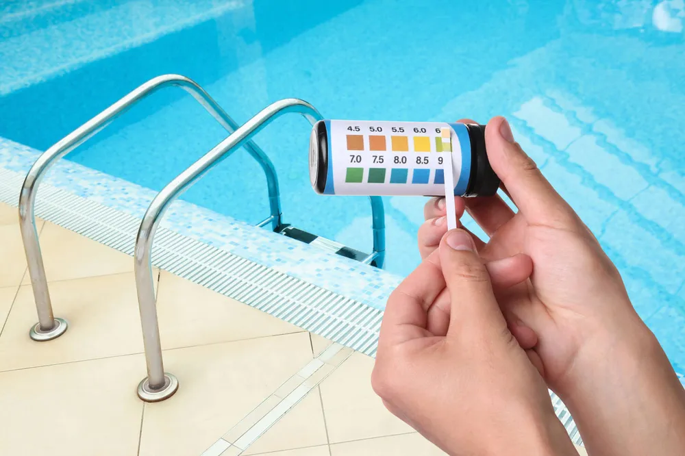 A pair of hands holding a pool water test strip against the printed colour chart on the side of its container, with the pool and its handrail behind