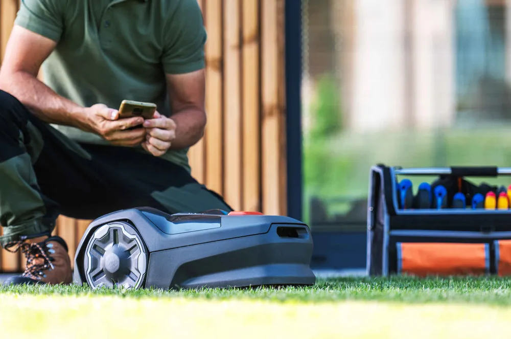A small autonomous robotic lawn mower sitting on a flat, closely mown lawn beside a kneeling person looking at a phone, with a toolbag on the grass