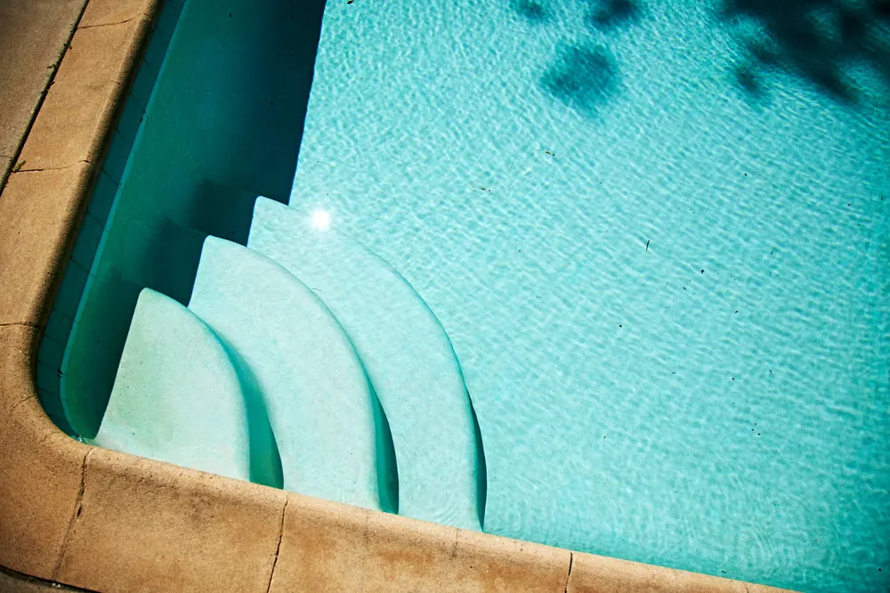 Three curved concrete steps in the corner of an outdoor swimming pool, seen from above through clear water beside warm sandstone coping
